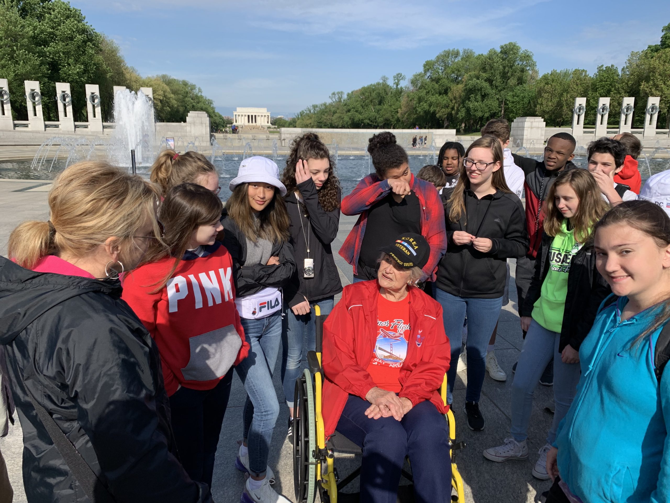 Group of middle school students standing around a woman US veteran in a wheelchair with Washington DC memorials in the background