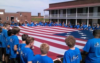 Adults and children in blue shirts holding large American flag - Lakeview Ft McHenry Flag Program