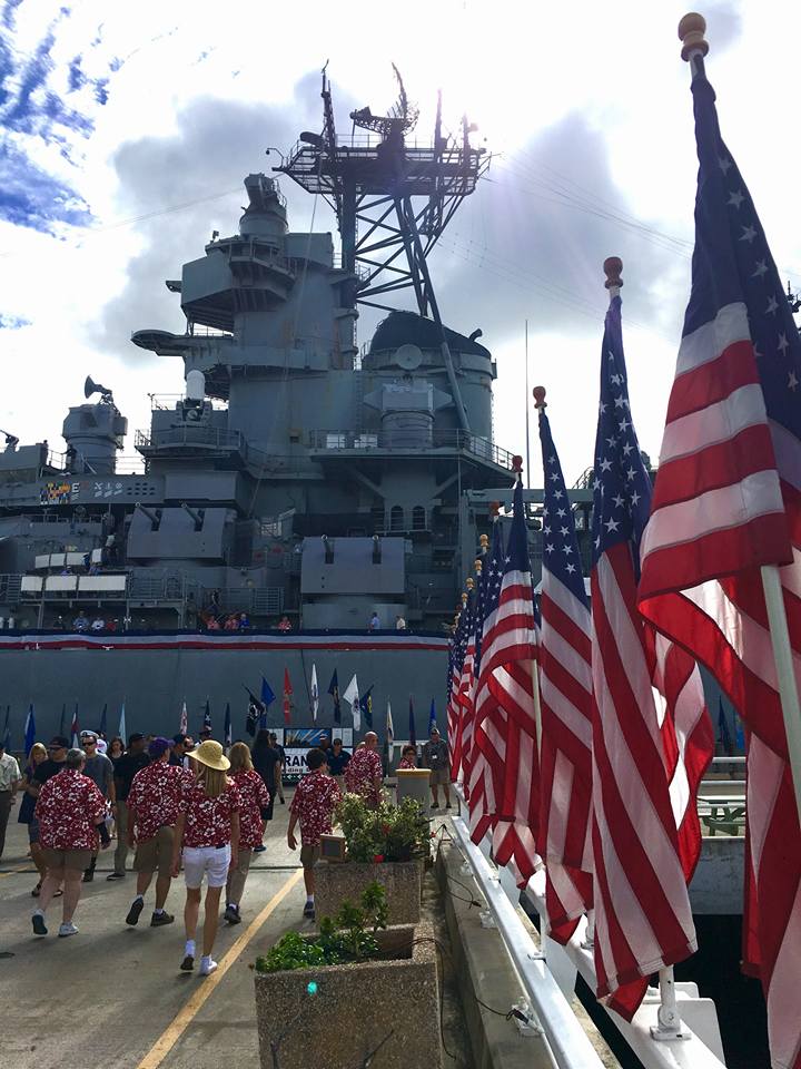 People walking on pearl harbor doc lined with American flags with historical battleship in the background